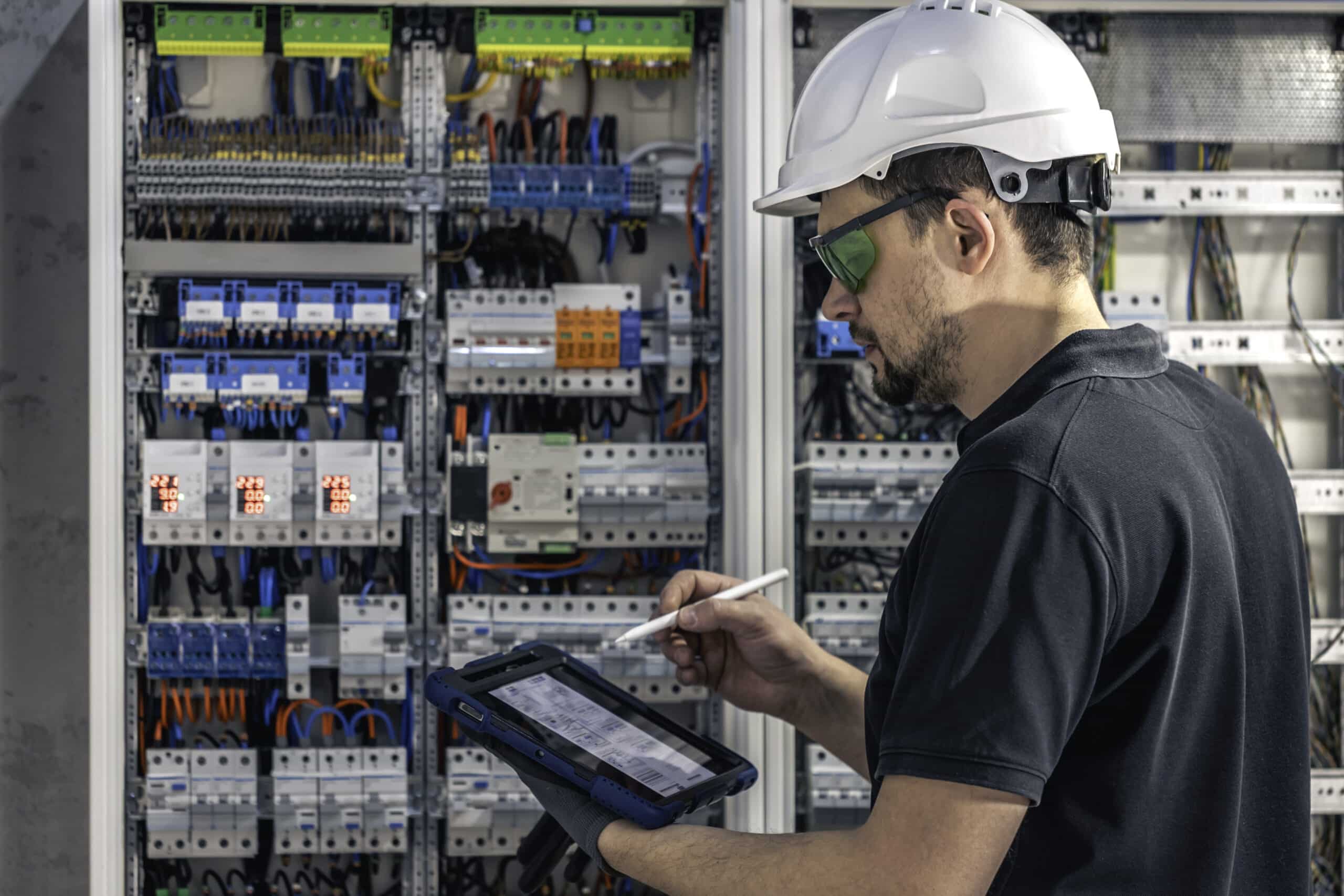 Man, an electrical technician working in a switchboard with fuses. Installation and connection of electrical equipment. Professional uses a tablet.